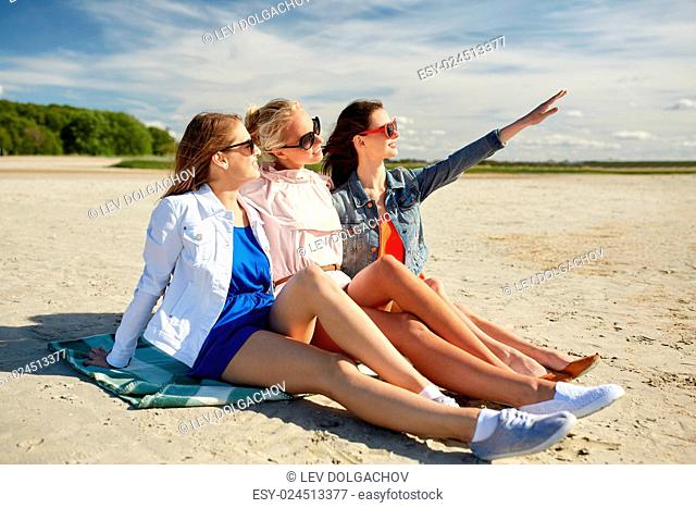 summer vacation, holidays, travel and people concept - group of smiling young women in sunglasses sitting on beach blanket and pointing finger to something