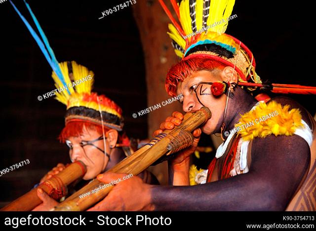 Two native indios in festive clothing and feathres in their hair playing their long pipe inside a hut, Mato Grosso, Brazil, South America