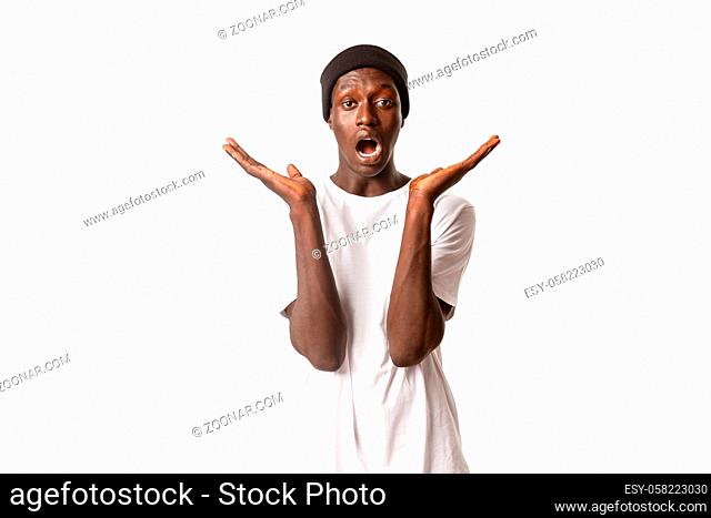 Portrait of surprised and confused handsome african-american male, open mouth and spread hands sideways amazed, standing white background in beanie and t-shirt