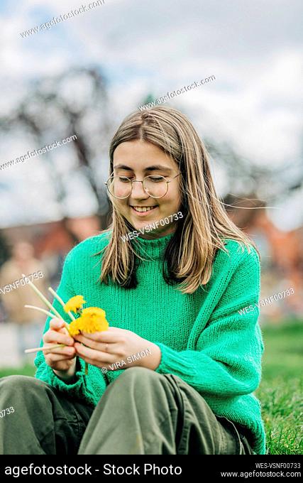 Smiling teenage girl making wreath with yellow dandelions sitting on grass