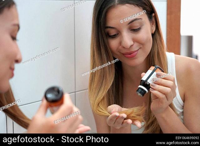 Close-up of young woman applying argan oil on hair tips in the bathroom at home. Hair care daily routine at home concept