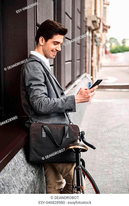 Smiling young businessman leaning on a wall while standing outdoors with a bicycle and using mobile phone