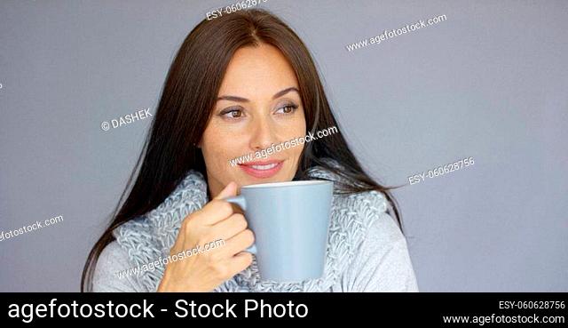 Beautiful long haired, brunette woman holding in her hand cup of hot drink. She wearing woolen scarf around her neck. She trying to warm up herself