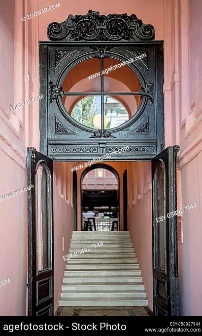 Entrance to the restaurant with dark wooden doors and a same round window with patterns. There are pink walls, stair to the hall with an arch door