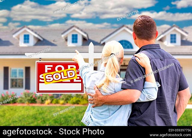 Young Adult Couple Facing and Pointing to Front of Sold Real Estate Sign and House