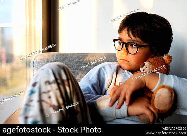 Portrait of serious boy with teddy bear sitting on armchair at home looking out of window