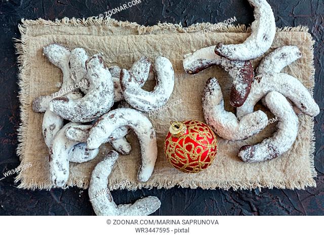 Homemade chocolate cookies in the form of a crescent sprinkled with powdered sugar on a linen cloth and a red Christmas ball on a textured dark background