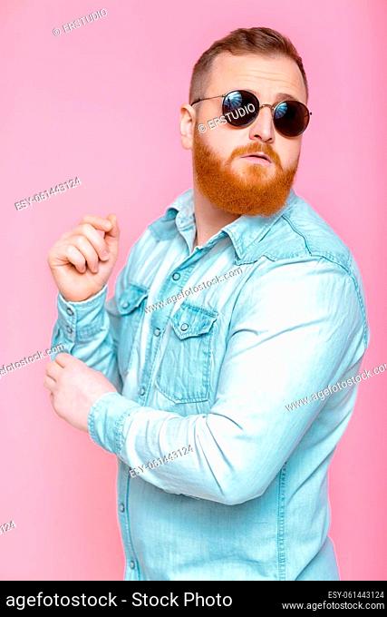 smiling red-haired beard man in sunglasses and denim shirt on pink background