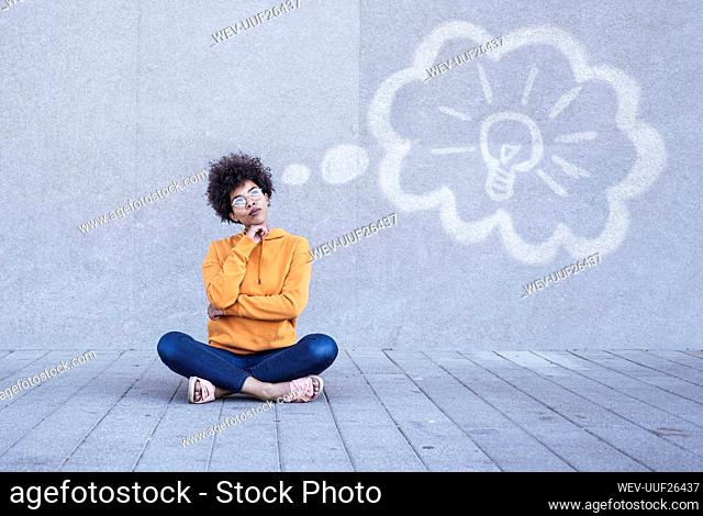 Pensive woman sitting on ground with electric bulb in thhought bubble