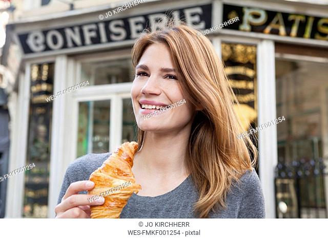 France, Paris, portrait of happy young woman with croissant in front of pastry shop