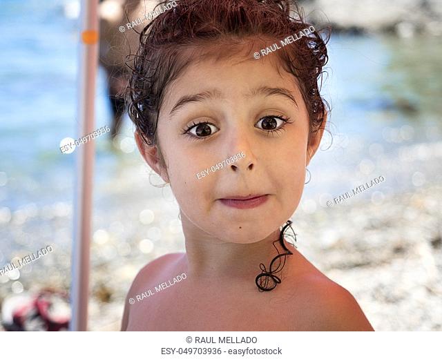 portrait of a kid mocking to the camera, she is shirtless on the beach, her curly hair is wet and she has it picked up, some curls hang down her neck