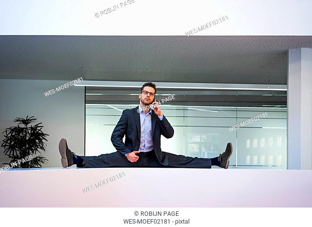 Businessman doing the splits on reception desk in office talking on cell phone