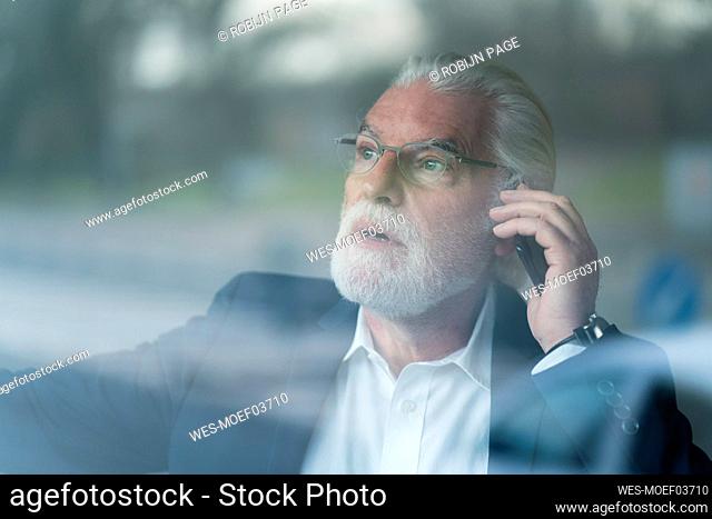 Surprised senior businessman talking on smart phone at office seen through glass