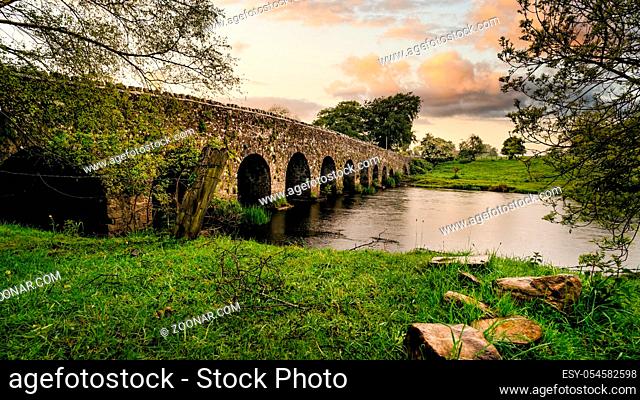 Old 12th century stone arch bridge over a river, rocks on first plane. Green fields and trees. Dramatic sky sunset. Count Meath, Ireland