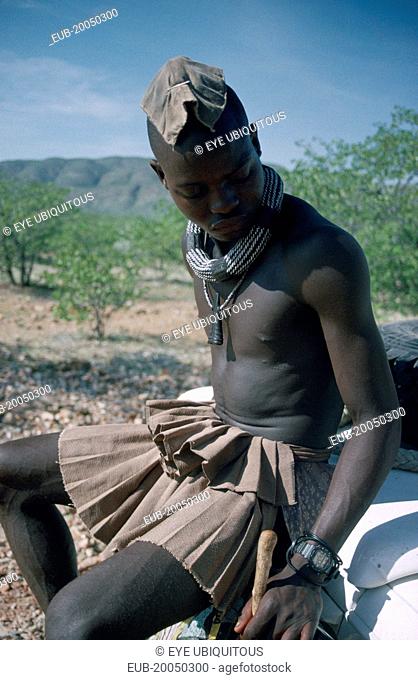 Kaokoland. Himba man sitting on bumper of safari jeep. Cloth covering head denotes that he is married