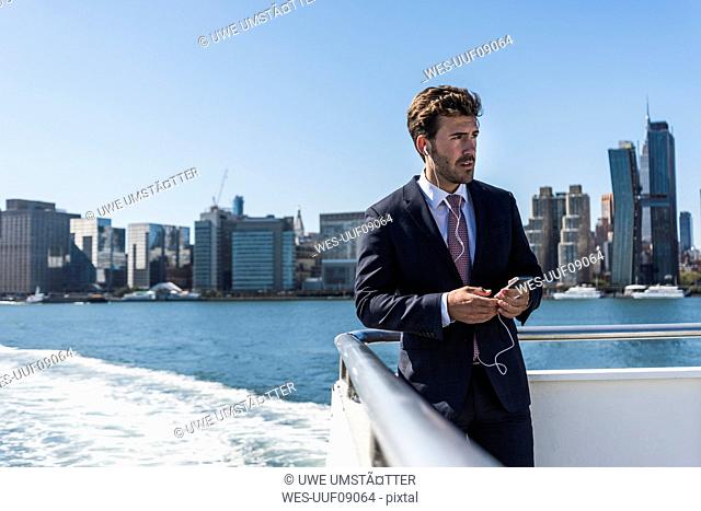 USA, New York City, businessman on ferry on East River with cell phone and earphones