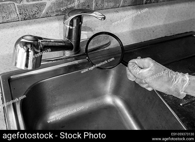 A close up and monochrome view of a person inspecting a kitchen sink and faucet with a magnification glass, checking for imperfections with copy space