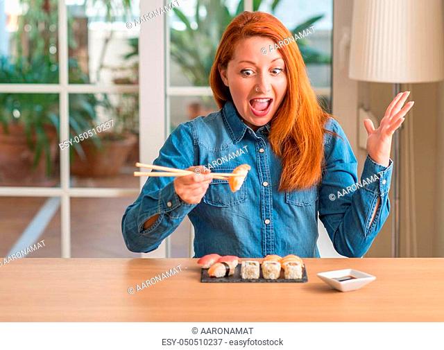 Redhead woman eating sushi using chopsticks very happy and excited, winner expression celebrating victory screaming with big smile and raised hands