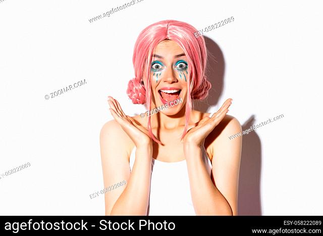 Close-up of attractive party girl in pink wig and bright makeup looking excited and smiling, standing over white background
