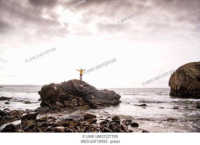 Rear view of woman with raised arms, standing at rocky beach