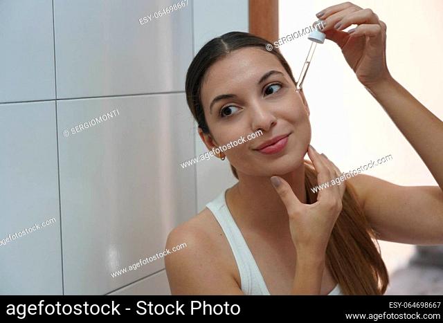 Beautiful young woman holding a pipette in her hand with serum moisturizing anti aging antioxidant looking in the mirror in her bathroom