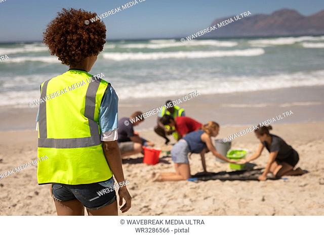 Rear view of mixed race female volunteer standing at beach while looking at the other volunteers on a sunny day