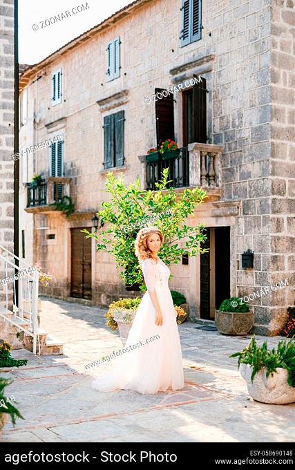 A gentle bride stands in a cozy courtyard near an old brick house withe beautiful balcony and windows with shutters. High quality photo