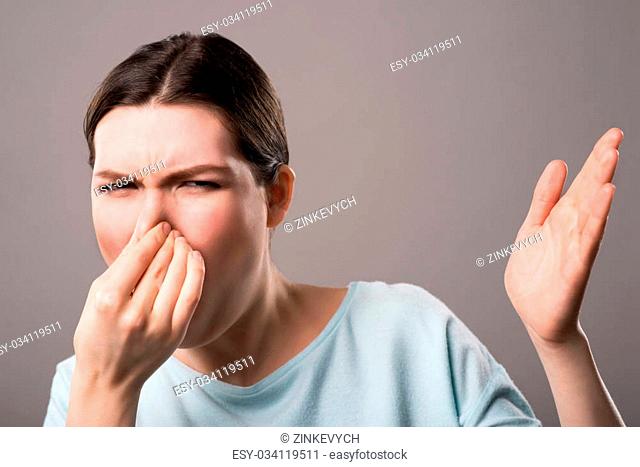 What a smell. Cheerless beautiful girl closing her nose and expressing disgust while standing isolated on grey background
