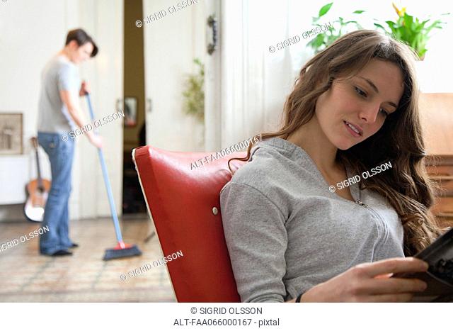 Woman reading magazine while her husband sweeps the floor