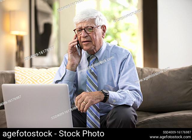 Mature Caucasian Male doctor practicing tele-medicine from his home, using cell phone and laptop computer, Talking to patient