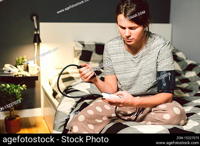 The topic of high blood pressure is hypertension disease. 22 year old young Caucasian woman uses an automatic tonometer to check pressure at home in the bedroom...