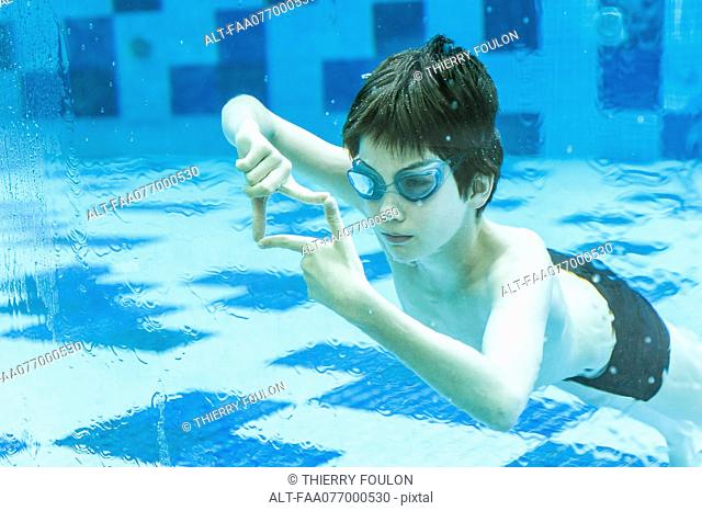 Boy wearing goggles swimming underwater in swimming pool, hands forming finger frame