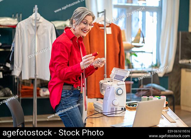 Workshop. Smiling stylish woman showing thread on laptop screen standing near table with sewing machine in atelier