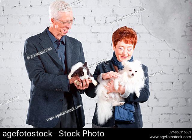 Happy smiling old couple standing together with pet rabbit and dog on white brick background