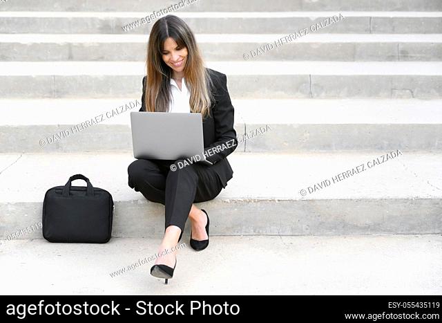 Beautiful businesswoman in suit, using a laptop sitting on stairs outdoors. High quality photo