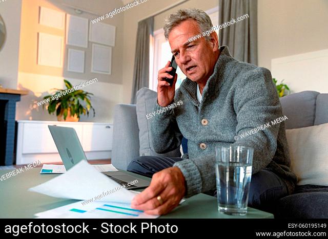 Focused caucasian senior man sitting on sofa, doing paperwork, making call and using laptop