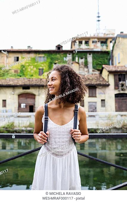 Italy, Milan, smiling young woman with backpack wearing white summer dress standing in front of water