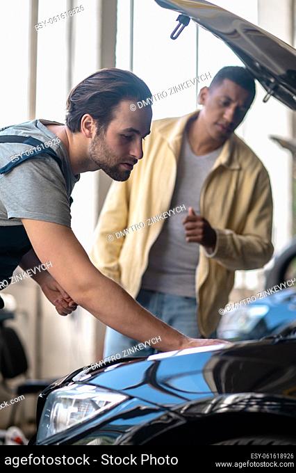 Finding problem. Young adult caucasian attentive mechanic identifying car problem touching insides of hood and excited dark-skinned client watching