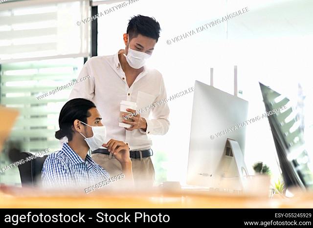 Two asian businessman talk discuss their work in morning after office reopen due to coronavirus COVID-19 pandemic. They wear protective face mask to prevent...