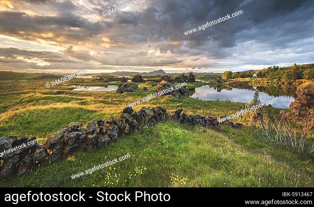 Stone wall and lava formations of volcanic rock rise from green meadow with dramatic sky in the evening light at Kalfaströnd