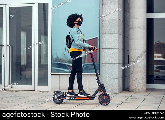 Mujer con mascarilla protectora montando scooter eléctrico por edificio durante la crisis de COVID-19