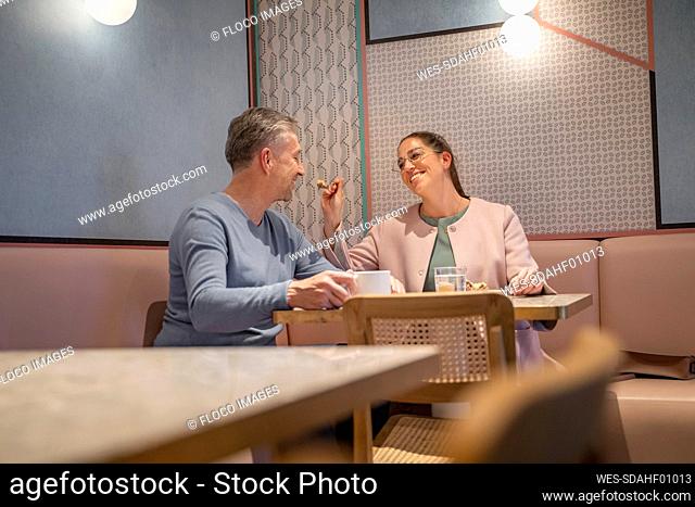 Smiling businesswoman feeding man while sitting at modern cafe