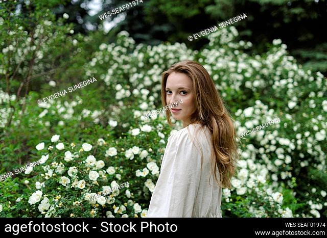 Young woman with long blond hair standing by white flower bush