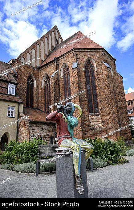 PRODUCTION - 20 July 2023, Brandenburg, Prenzlau: A harlequin figure sits making music in front of the former Dominican monastery in Prenzlau