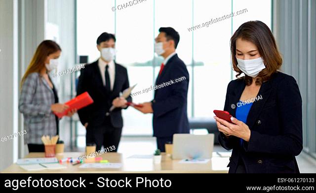 An Asian businesswoman in suit standing with mobile phone in her hand. The team is preparing for the meeting in the background