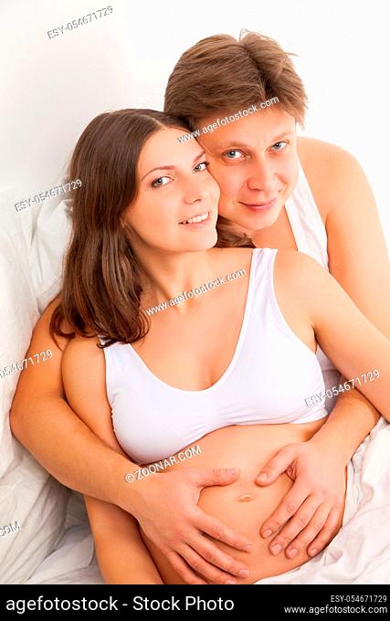 happy young pregnant woman with husband on white bed in light bedroom