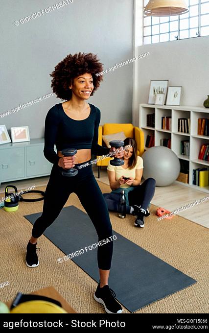 Smiling woman exercising with dumbbell by friend using smart phone in living room