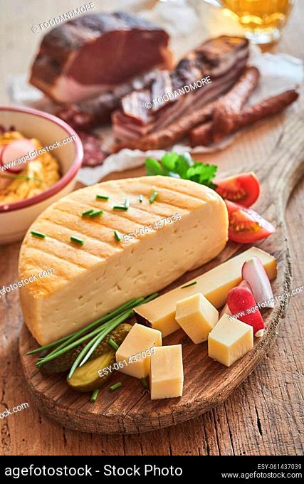 Delicious cheese with cubes decorated with green onion served on wooden chopping board on table with sausages and smoked lard in kitchen