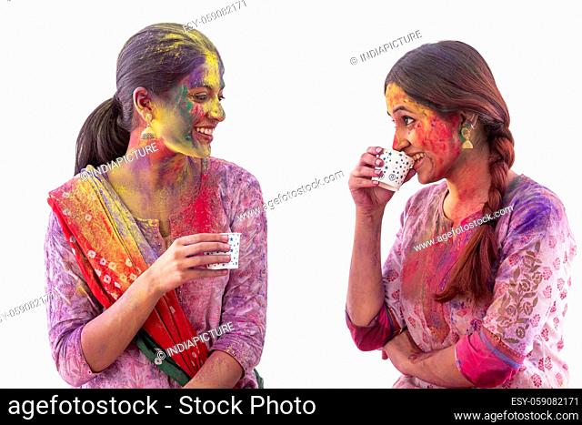 TWO YOUNG WOMEN WITH COLOURS ALL OVER HAPPILY LOOKING AT EACH OTHER AND DRINKING TEA