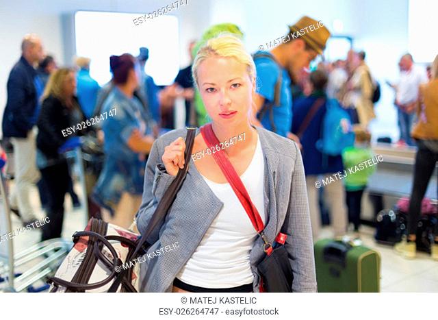 Casually dressed young stylish female traveller waiting in airport arrival terminal hall, collecting her laggage . Blured background